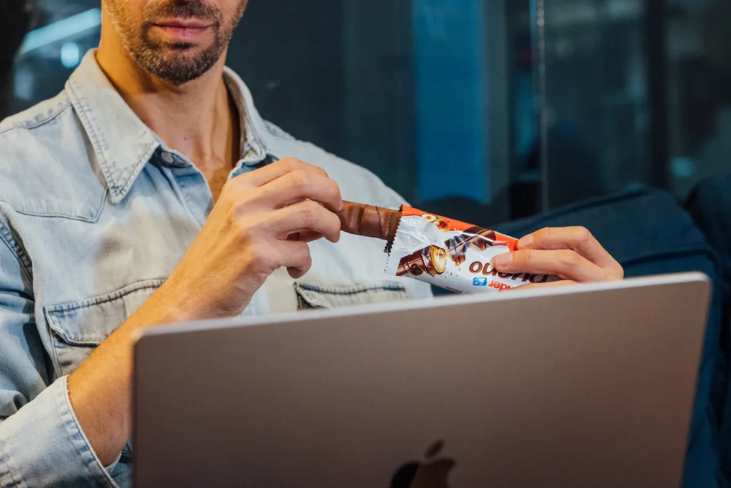 Profesional comiendo una barrita de chocolate mientras trabaja en su portátil durante la pausa de media mañana en la oficina