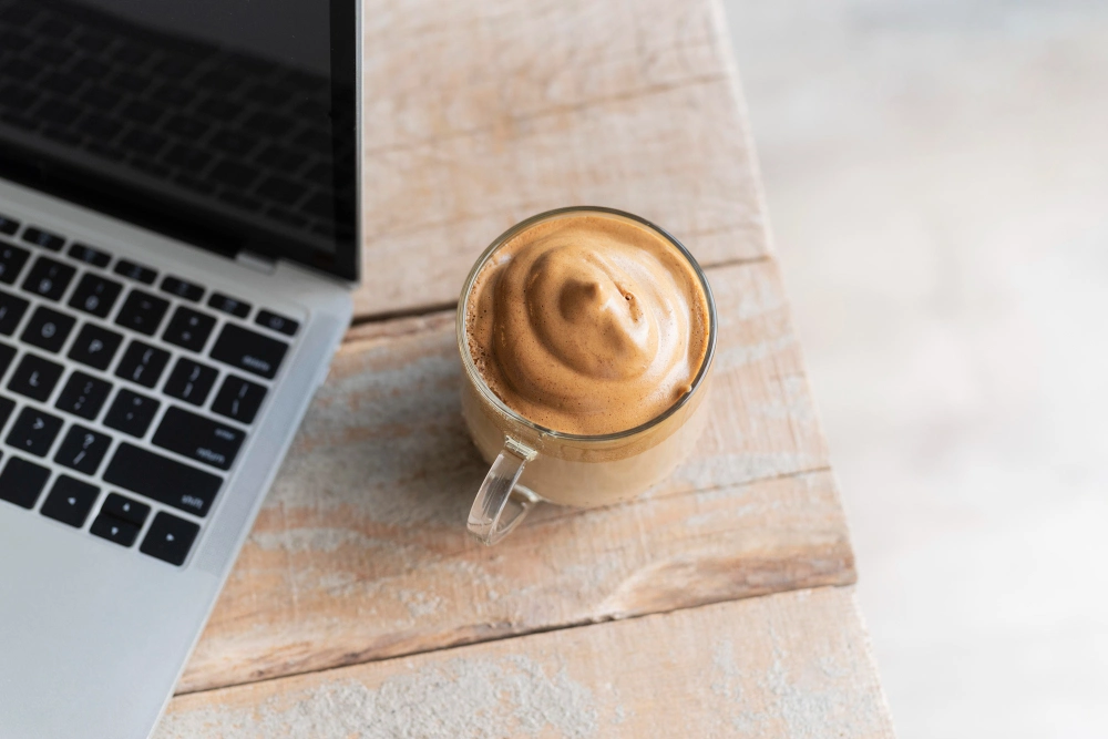 Taza de café mocha cremoso junto a un portátil en una mesa de trabajo de oficina.