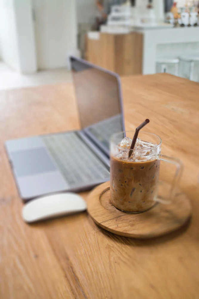 Café frío con hielo servido en vaso junto a un ordenador portátil en una mesa de trabajo.